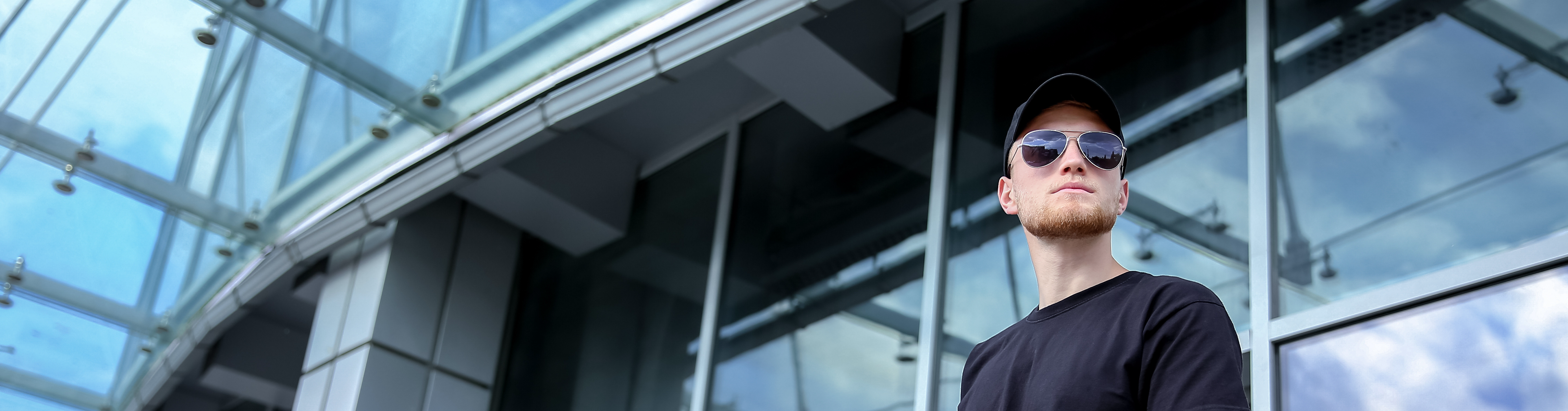 A figure in dark navy clothing and sunglasses stands below a glass and steel building, scanning the surroundings