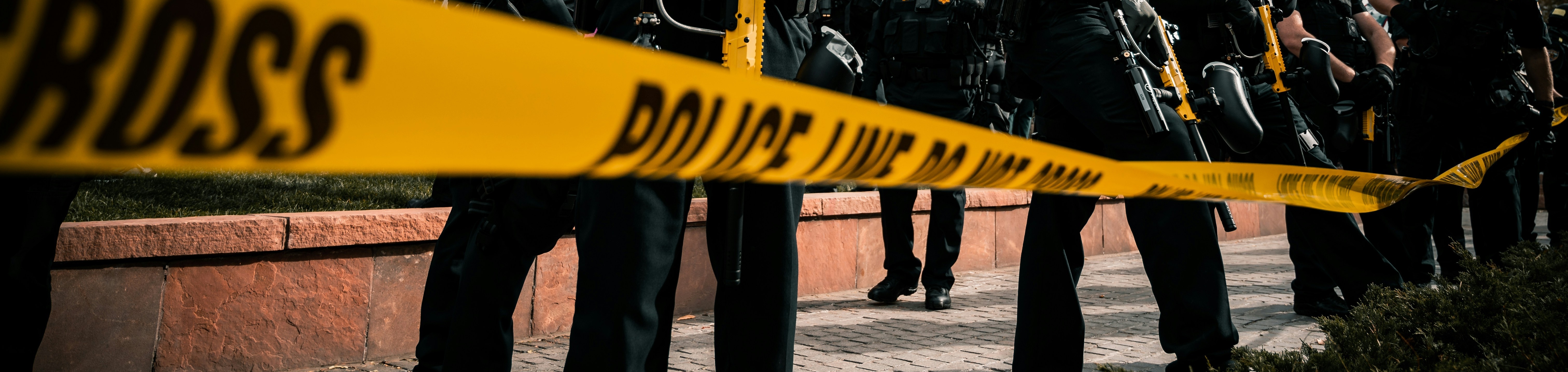 Yellow police tape reading Police Line Do Not Cross stretches across a scene where officers in tactical gear stand beyond the cordon